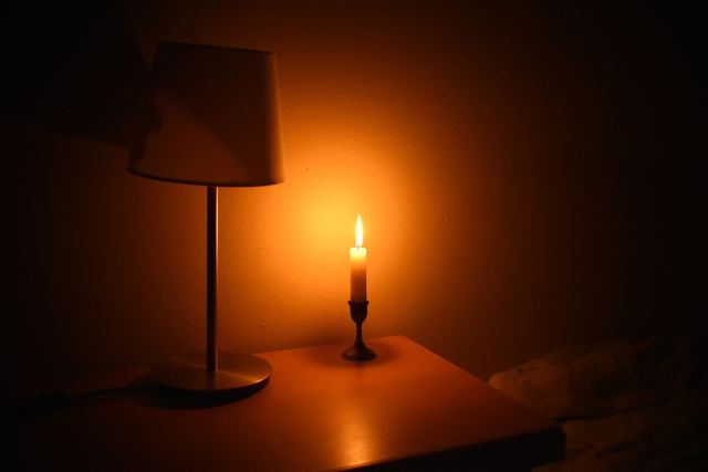 A bedside table in a dark room with a table lamp switched off and a single lit candle beside it.