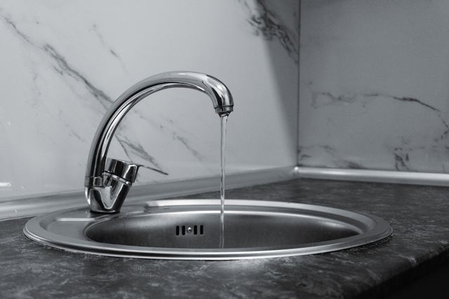 Black and white photo of a chrome kitchen mixer tap running water into a round stainless steel sink.