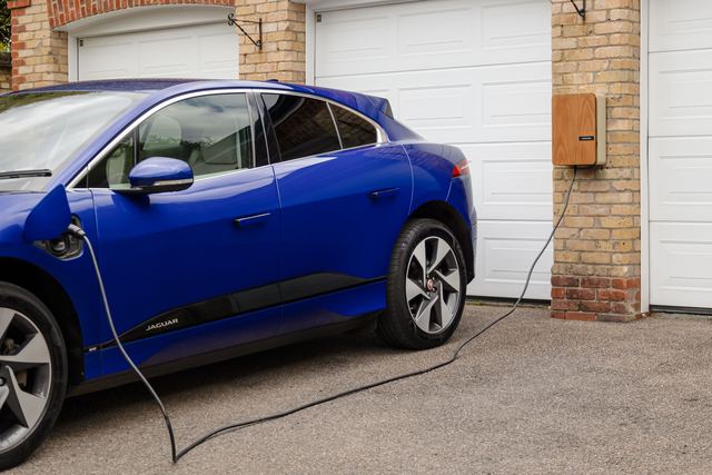 A blue electric car on a driveway plugged into a wall-mounted charger on a brick garage pillar.