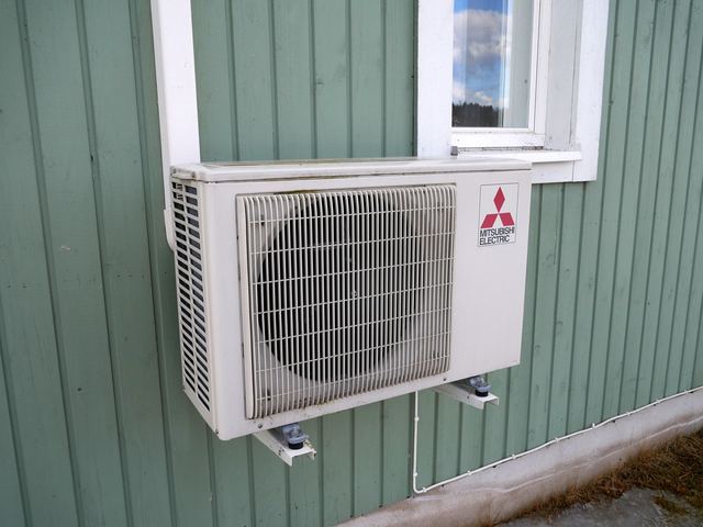 A heat pump outdoor unit on wall brackets against green weatherboards, with a white cable conduit running down the wall below it.