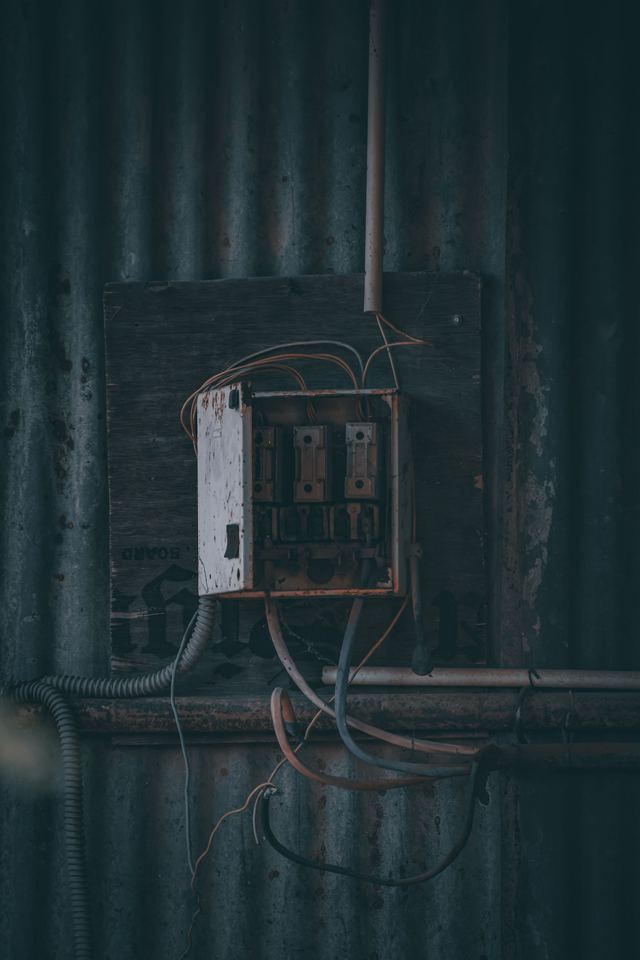 An old rusty fuse box with three ceramic rewireable fuses on a dark board, with loose wires against a corrugated iron wall.