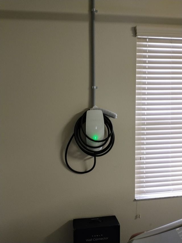 A Tesla Wall Connector charger with its cable coiled around it, mounted on a garage wall with conduit running to the ceiling.